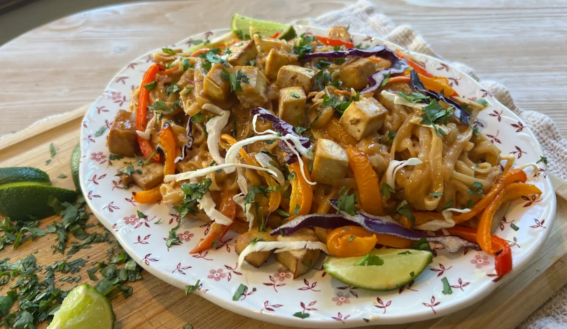 A plate of stir-fried noodles with tofu, sliced bell peppers, red cabbage, and fresh herbs, garnished with lime wedges.