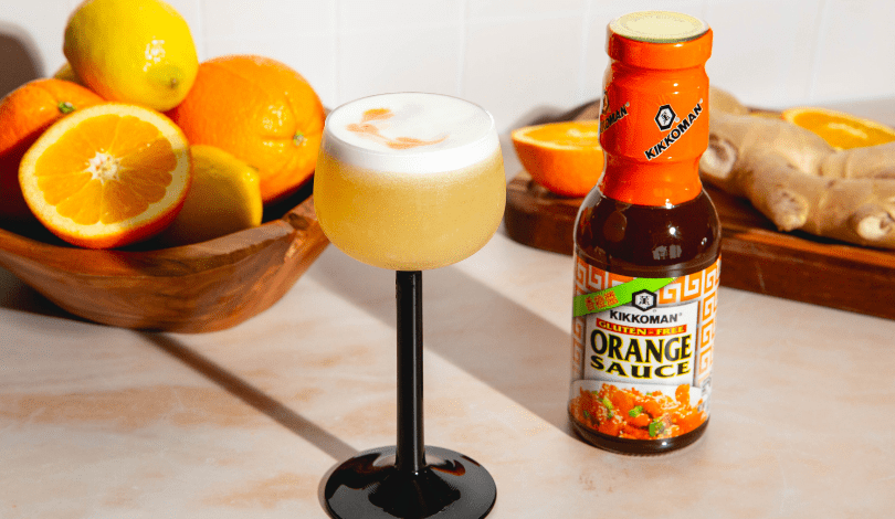 A cocktail with a foamy top in a black-stemmed glass sits next to a bottle of Kikkoman Orange Sauce, with fresh oranges and ginger in the background.