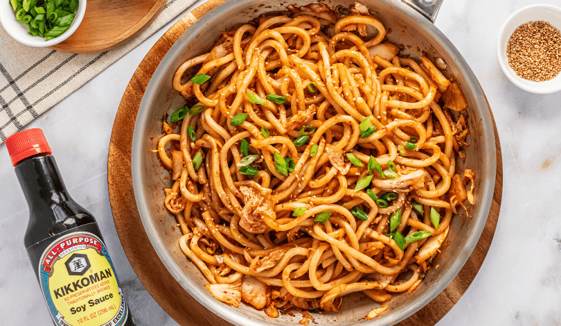 A pan of stir-fried udon noodles with vegetables, garnished with sliced green onions, next to a bottle of soy sauce and small bowls of sesame seeds and green onions.