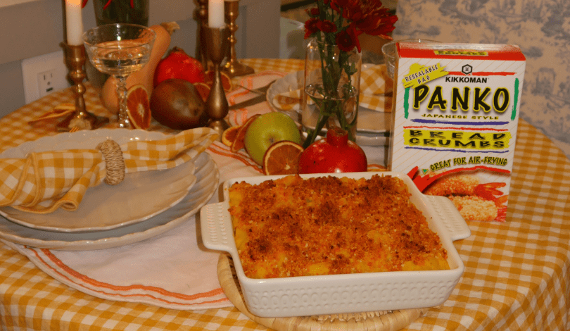 A table set with plates, utensils, a napkin, candles, fruit, and flowers. A dish of baked macaroni and cheese with a box of Panko bread crumbs is in the foreground.