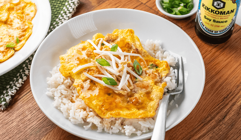 A plate of white rice topped with an egg omelet, bean sprouts, and green onions, with a fork on the plate. A small bowl of green onions and a bottle of soy sauce are nearby.