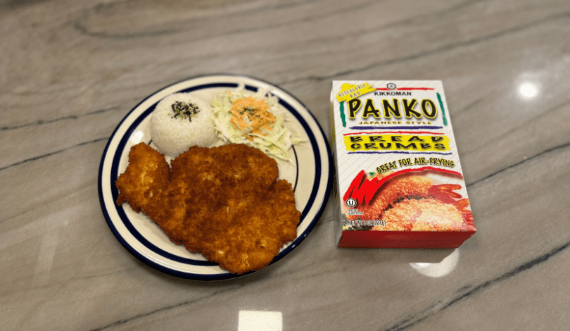 A plate with breaded fried cutlet, white rice with seasoning, and shredded cabbage beside a box of Kikkoman Panko bread crumbs on a countertop.