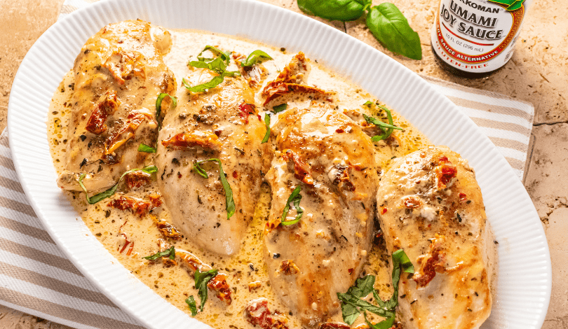 Four baked chicken breasts in a creamy sauce with herbs and sun-dried tomatoes, served in a white oval dish on a striped towel. A bottle of seasoning sauce is visible in the background.