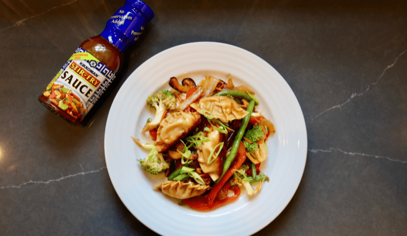 A plate of stir-fried vegetables and dumplings garnished with green onions next to a bottle of stir fry sauce on a gray surface.