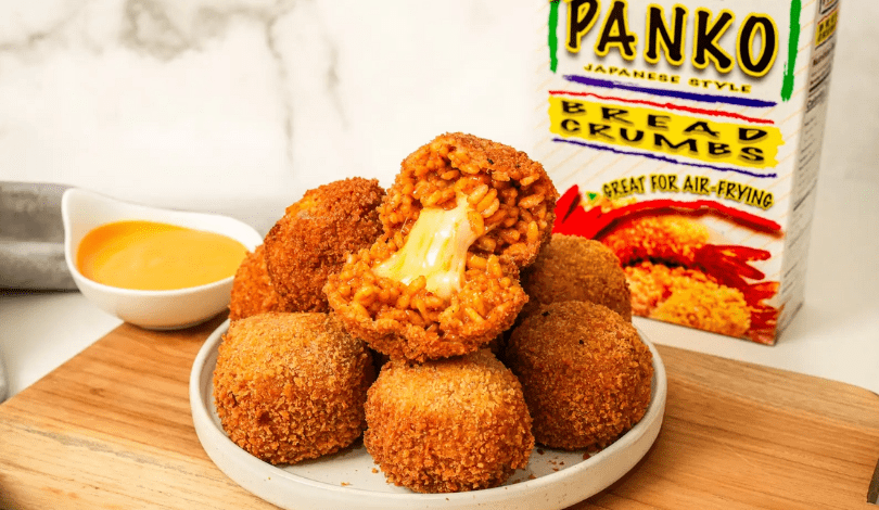 A plate of breaded, fried balls filled with macaroni and cheese, with a box of panko bread crumbs and a bowl of dipping sauce in the background.
