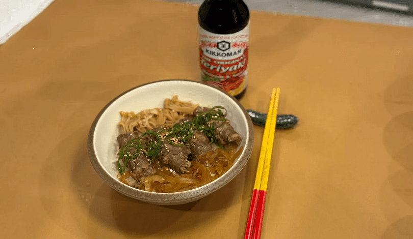 A bowl of noodles with meat, green onions, and sauce, next to yellow and red chopsticks, a bottle of Kikkoman teriyaki sauce, and a small cucumber on a brown surface.
