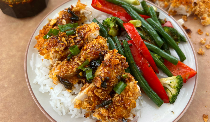 A plate of white rice topped with breaded chicken, garnished with green onions, served with sautéed green beans, red bell peppers, and broccoli.