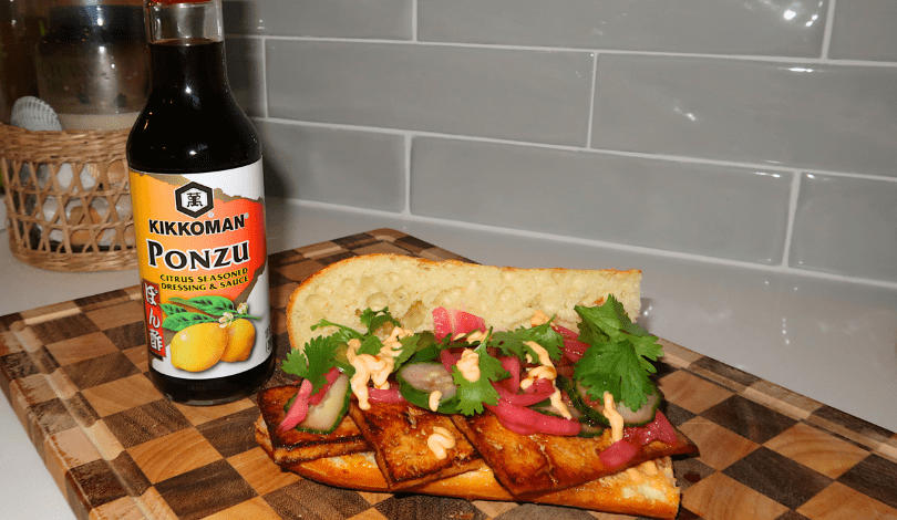 A bottle of Kikkoman Ponzu sauce next to a sandwich with tofu, pickled onions, cucumbers, cilantro, and sauce on a wooden cutting board.