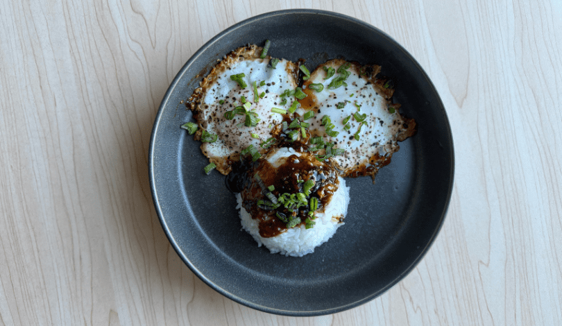 A black plate with two fried eggs topped with chopped green onions and ground pepper, served alongside a mound of white rice drizzled with a dark sauce.