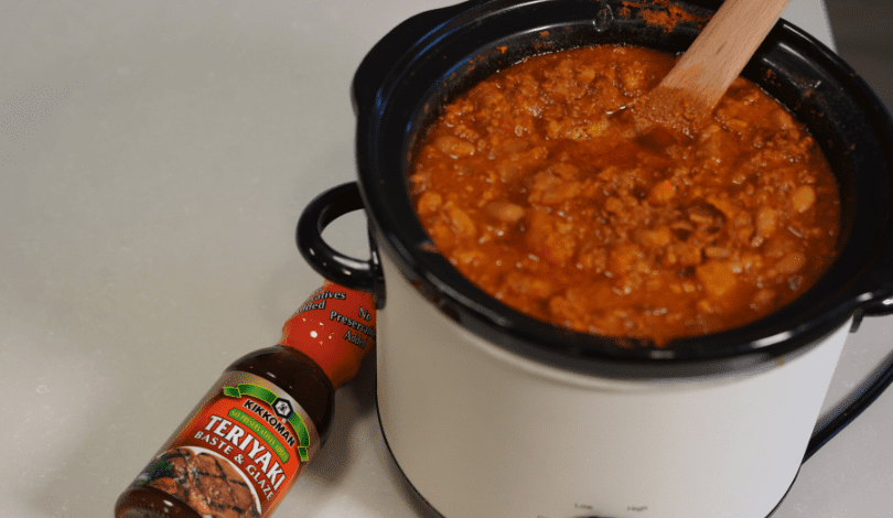 A slow cooker filled with a thick orange-red stew sits on a counter beside a bottle of Kikkoman Teriyaki marinade and sauce.