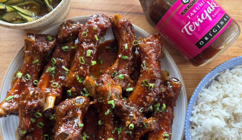 A plate of glazed ribs garnished with chopped green onions, served with white rice, sliced cucumbers, and a bottle of teriyaki sauce.