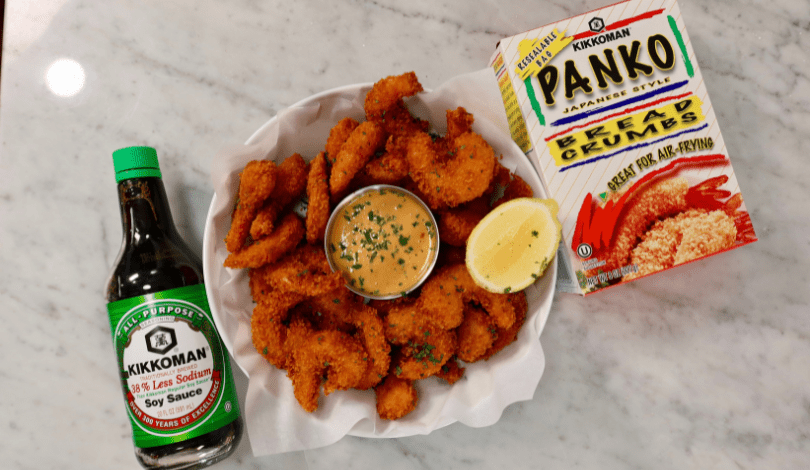 A plate of breaded fried shrimp with dipping sauce and a lemon wedge, next to a bottle of Kikkoman soy sauce and a box of Kikkoman panko bread crumbs on a marble surface.