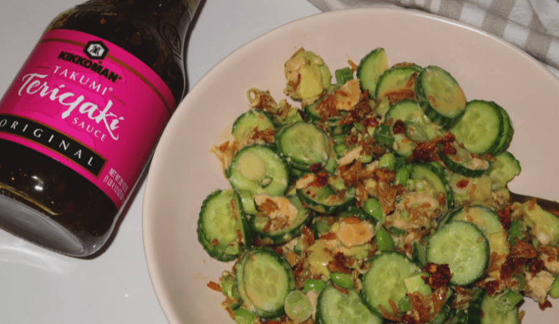 A bowl of cucumber salad with edamame and crispy toppings beside a bottle of Kikkoman Takumi Teriyaki Sauce.