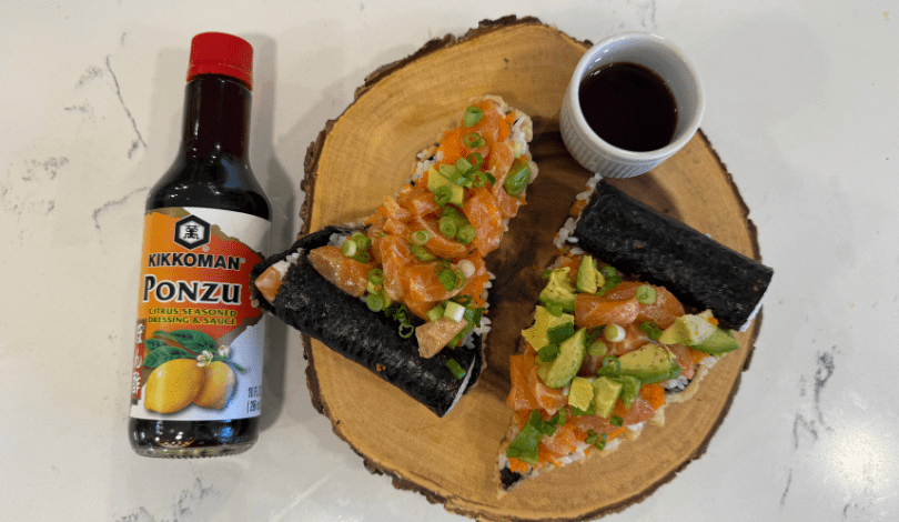 Three hand-rolled sushi cones with salmon, avocado, and green onions are arranged on a wooden board beside a bottle of Kikkoman Ponzu sauce and a small dish of sauce.