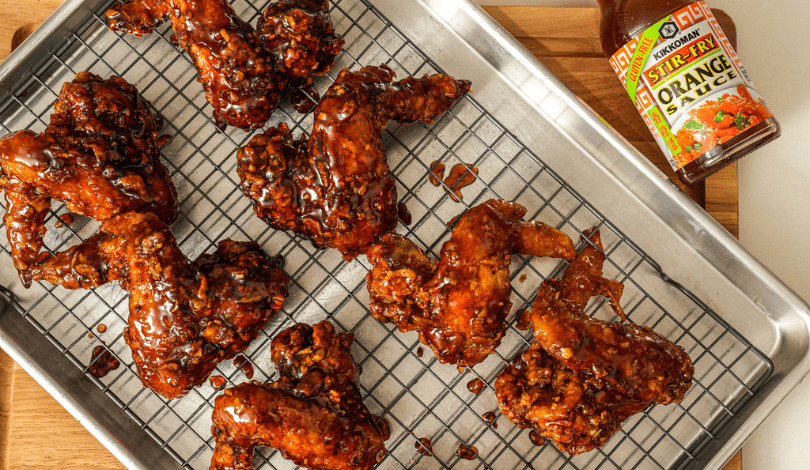 Chicken wings coated in a glossy orange glaze rest on a wire rack over a baking sheet, with a bottle of stir-fry orange sauce nearby.
