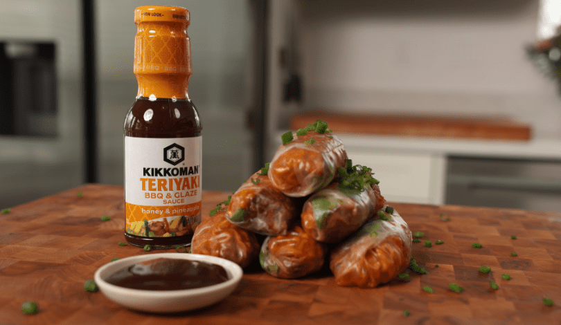 A bottle of Kikkoman Teriyaki BBQ & Glaze sauce next to a stack of fresh spring rolls, garnished with chopped herbs, and a small dish of sauce on a wooden surface.