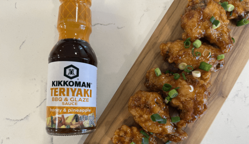 A bottle of Kikkoman Teriyaki BBQ & Glaze Sauce next to glazed chicken wings garnished with chopped green onions on a wooden board.