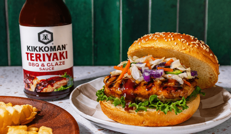 A chicken burger with lettuce and coleslaw sits on a plate next to a bottle of Kikkoman Teriyaki BBQ & Glaze sauce and a side of crinkle-cut fries.