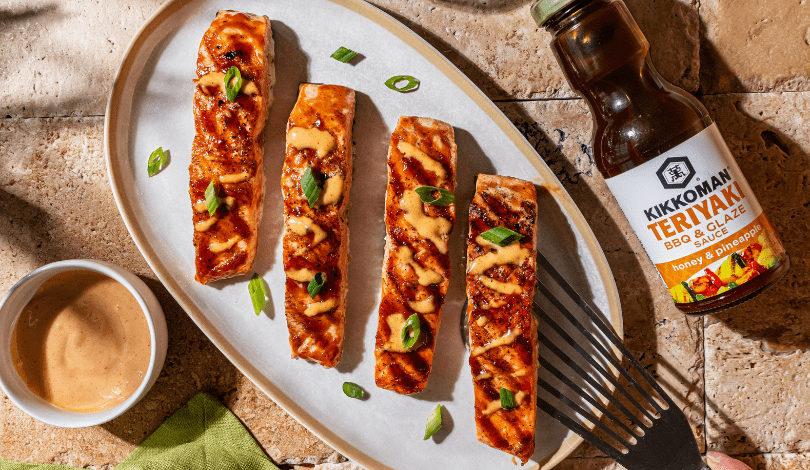 Four grilled salmon fillets on a white plate, drizzled with sauce and garnished with green onions, next to a bottle of Kikkoman Teriyaki BBQ glaze and a small bowl of dipping sauce.