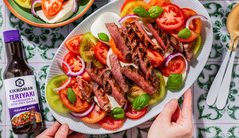 A platter of grilled steak slices on a bed of colorful tomato and red onion salad, garnished with basil, next to a bottle of Kikkoman teriyaki sauce.