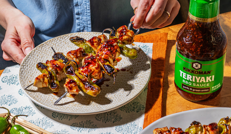 A person places grilled chicken and vegetable skewers on a plate, next to a bottle of Kikkoman Teriyaki BBQ Sauce on a wooden table.