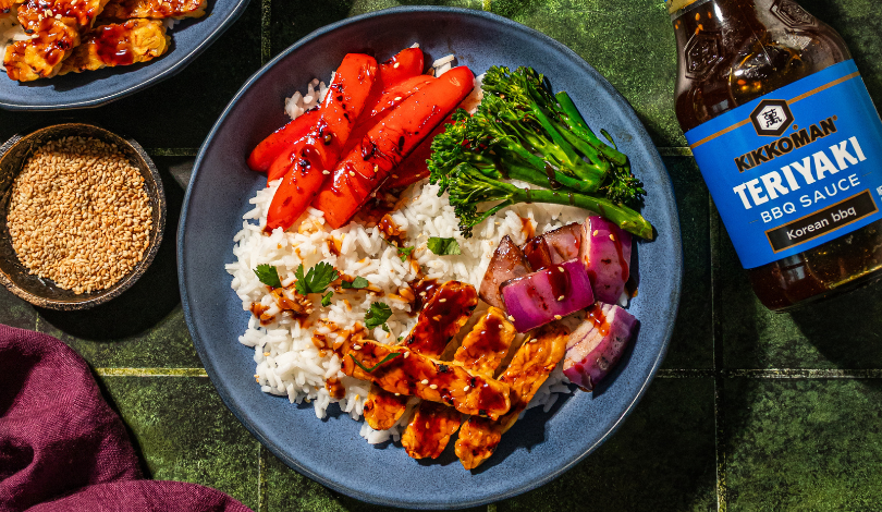 A bowl with white rice, sliced carrots, broccolini, red onion, and glazed tempeh, garnished with herbs, next to a bottle of Kikkoman Teriyaki BBQ Sauce and a dish of sesame seeds.