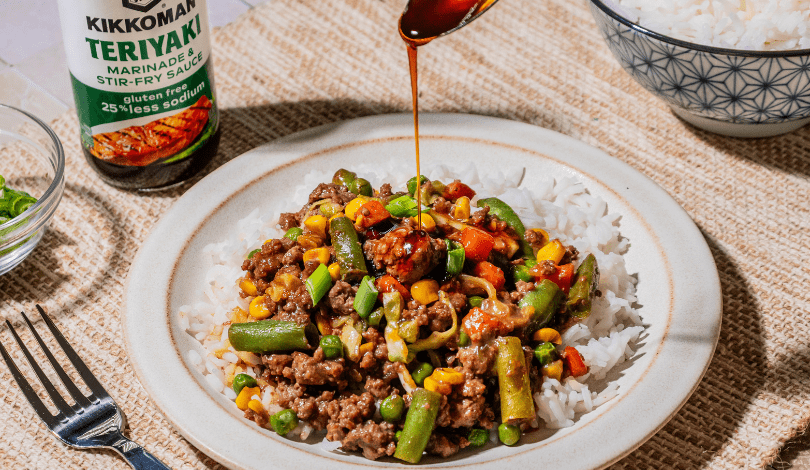 A plate of white rice topped with stir-fried ground beef and mixed vegetables, with teriyaki sauce being poured over it. A bottle of Kikkoman Teriyaki Sauce is nearby.
