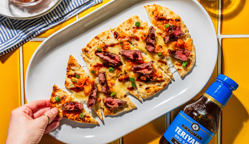 A hand takes a slice of flatbread topped with steak, cheese, bacon, and scallions on a white platter next to a bottle of Kikkoman Teriyaki BBQ sauce.