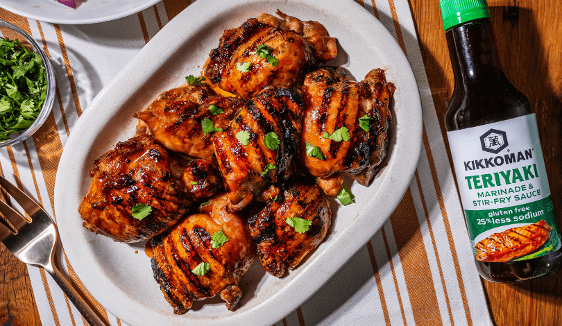 Grilled chicken thighs garnished with cilantro on a white plate, next to a bottle of Kikkoman teriyaki marinade and a small bowl of chopped herbs.