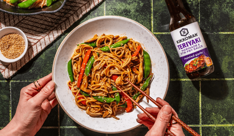 A person uses chopsticks to eat a bowl of stir-fried noodles with vegetables. A bottle of Kikkoman teriyaki sauce and a bowl of sesame seeds are nearby on a green tiled surface.