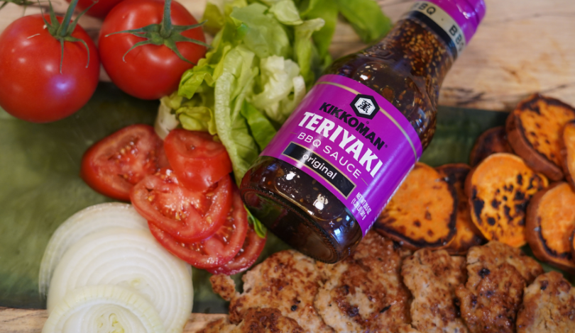 A bottle of Kikkoman Teriyaki BBQ Sauce on a table with sliced tomatoes, onions, lettuce, grilled chicken, and roasted sweet potatoes.
