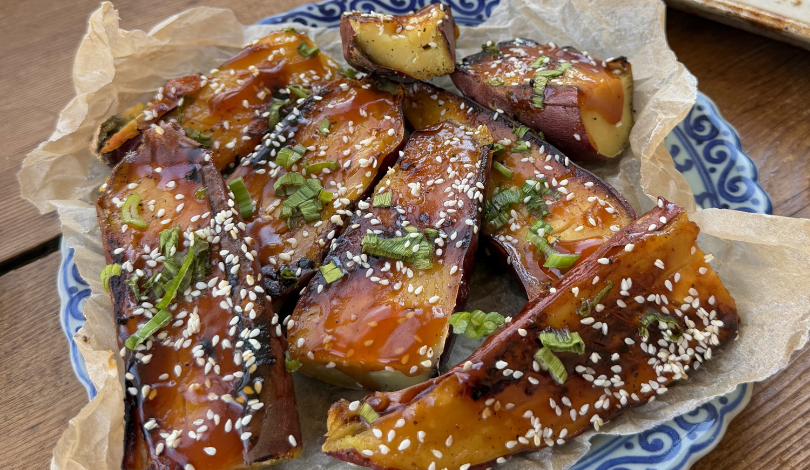 Slices of roasted sweet potato topped with a glossy sauce, chopped green herbs, and white sesame seeds, served on parchment paper in a blue and white patterned dish.