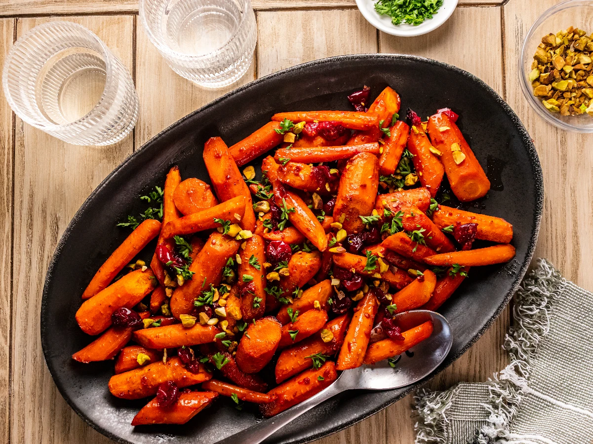 Oval black plate with roasted carrot pieces, dried cranberries, chopped pistachios, and herbs, on a wooden table; two empty glasses and small bowls nearby.