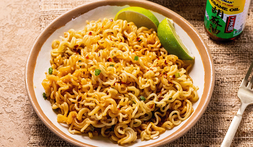 A plate of cooked instant noodles garnished with red pepper flakes and green onions, served with lime wedges. A bottle of sesame oil is visible in the background.