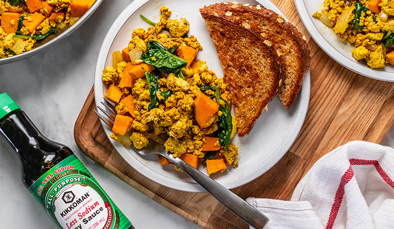 A plate with tofu scramble, sautéed vegetables, and two slices of toasted bread. A bottle of Kikkoman soy sauce and a towel are nearby.