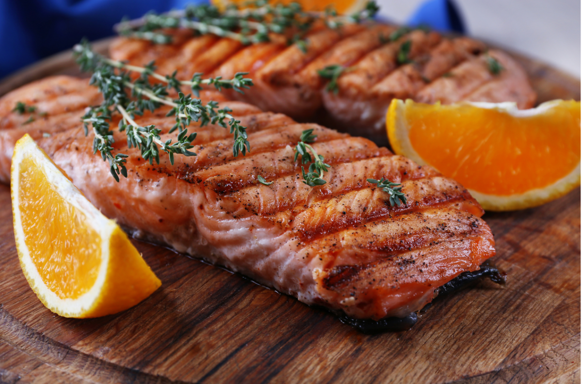Grilled salmon fillets on a wooden board, garnished with sprigs of fresh herbs and served with orange wedges.