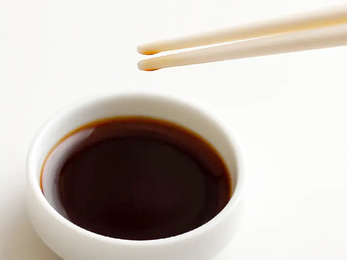 A pair of chopsticks held above a small white bowl filled with dark soy sauce on a plain white background.