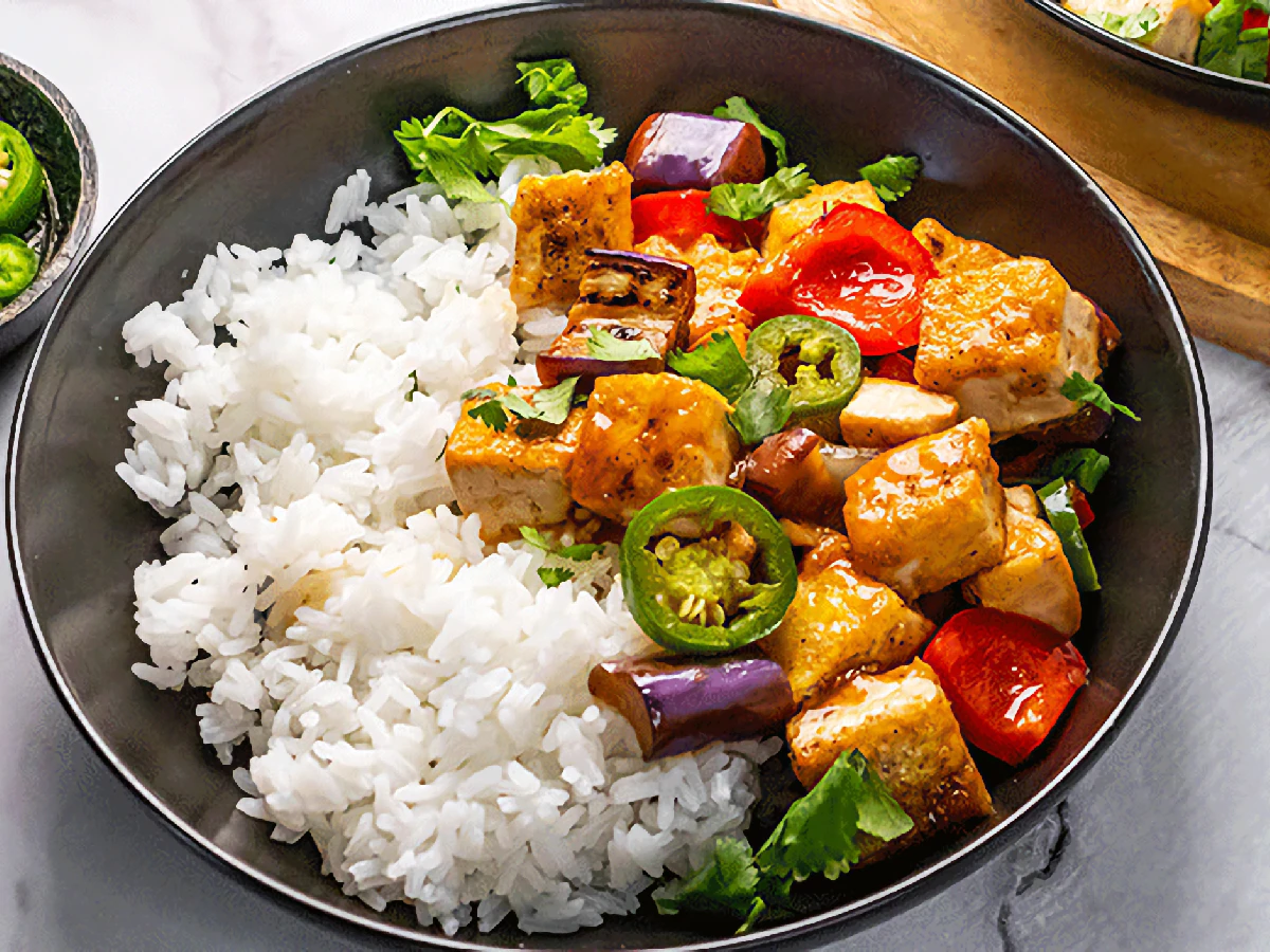 A bowl of white rice served with sautéed tofu, eggplant, red bell pepper, jalapeño slices, and fresh cilantro.