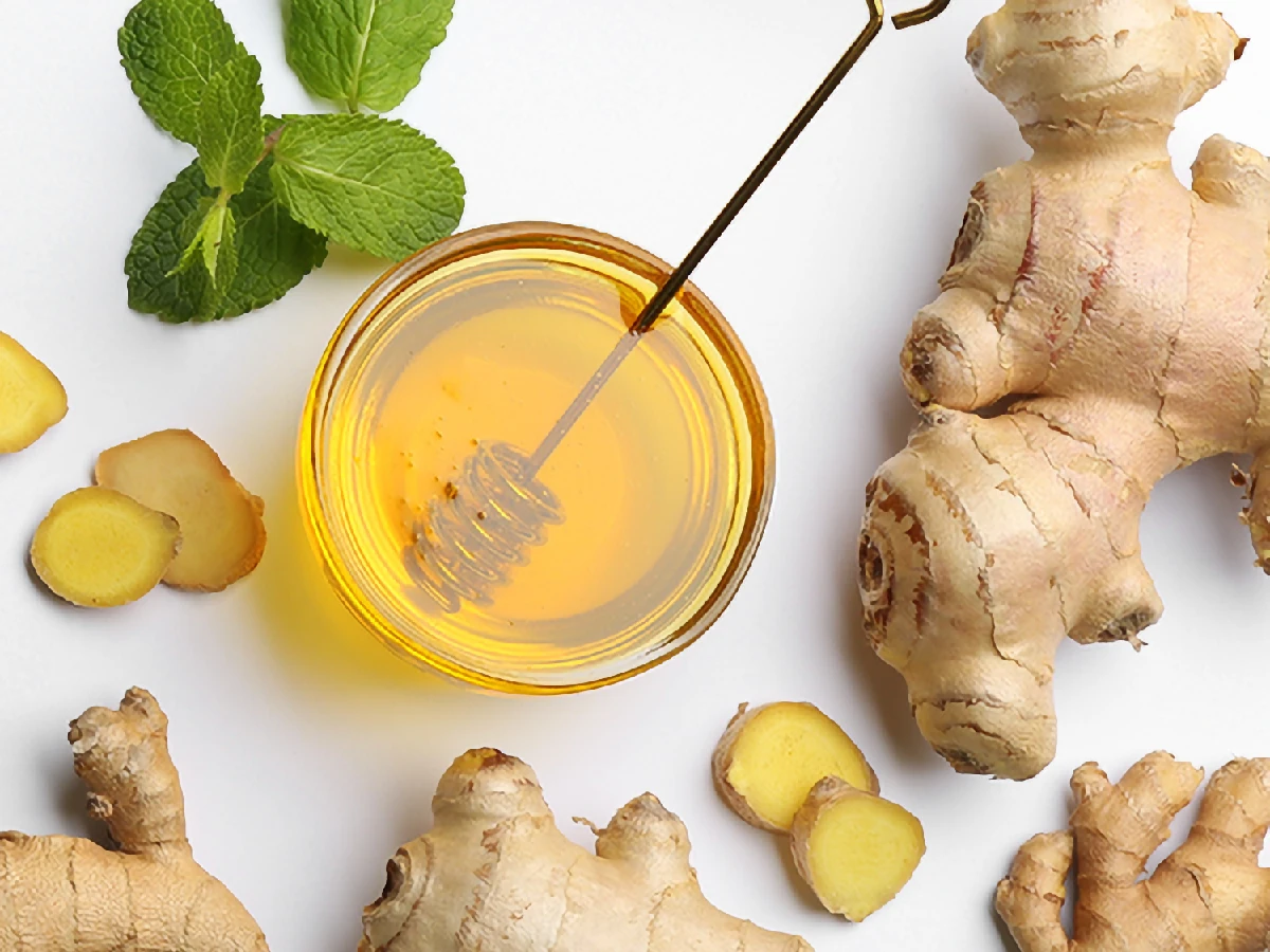A bowl of honey with a dipper, surrounded by fresh ginger roots, ginger slices, and a sprig of mint on a white background.
