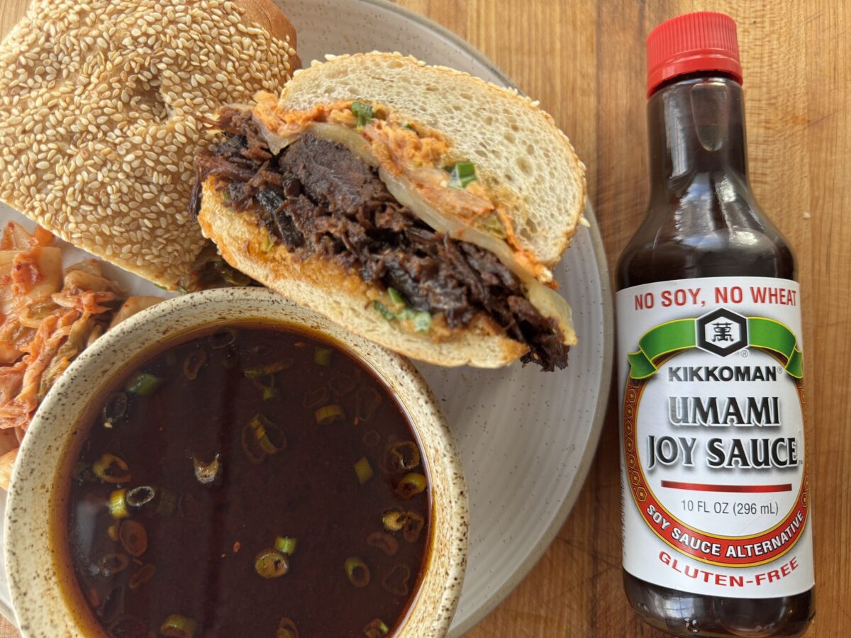 A sandwich with shredded meat and kimchi on a sesame bun, a bowl of broth with green onions, kimchi on the side, and a bottle of Kikkoman Umami Joy Sauce.