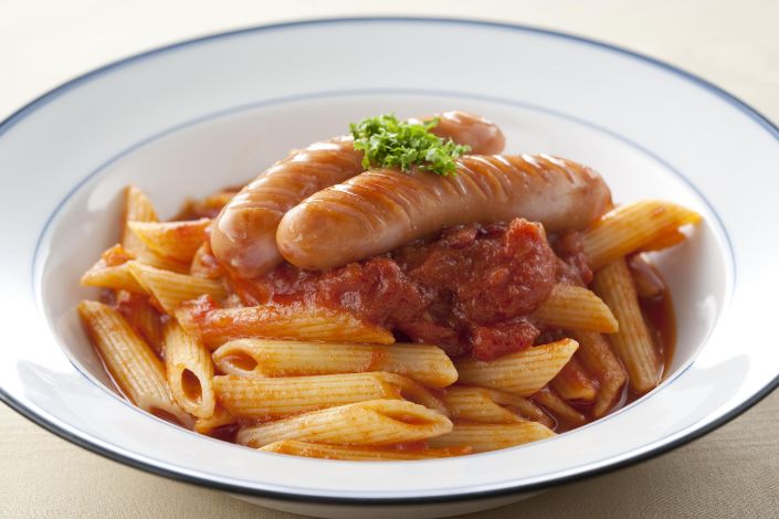 A white bowl containing penne pasta in tomato sauce, topped with two sausages and a garnish of chopped herbs.