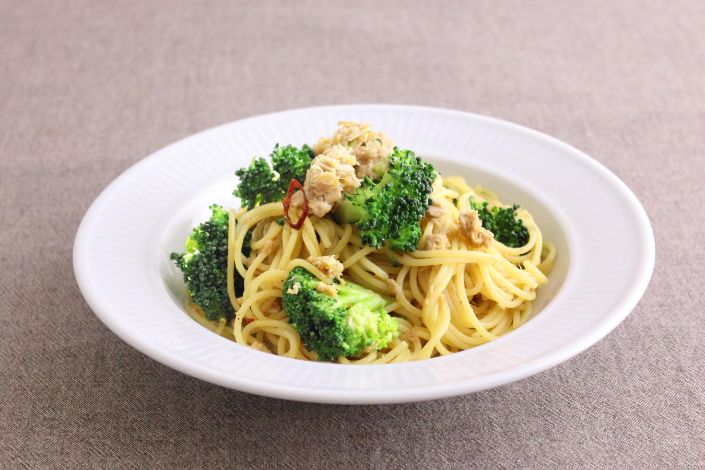 A white plate of spaghetti with broccoli florets, ground meat, and red chili slices on a plain, light-colored surface.
