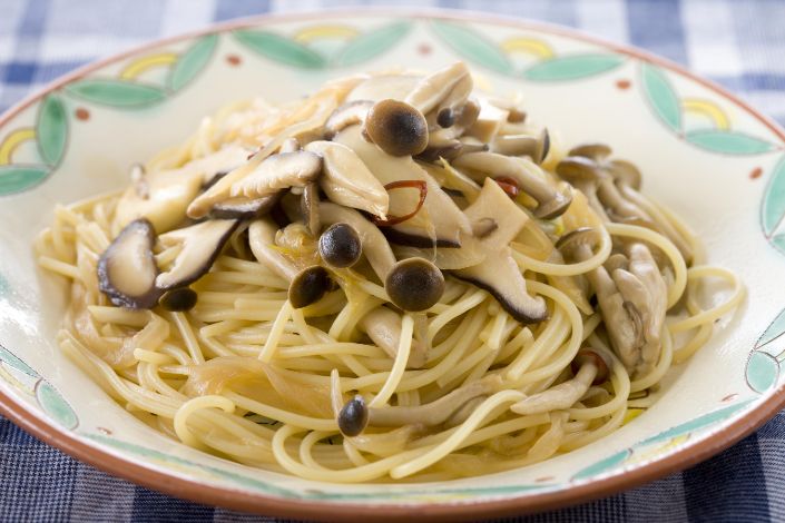 A plate of spaghetti topped with a mix of sliced mushrooms, served on a patterned dish over a blue checkered tablecloth.
