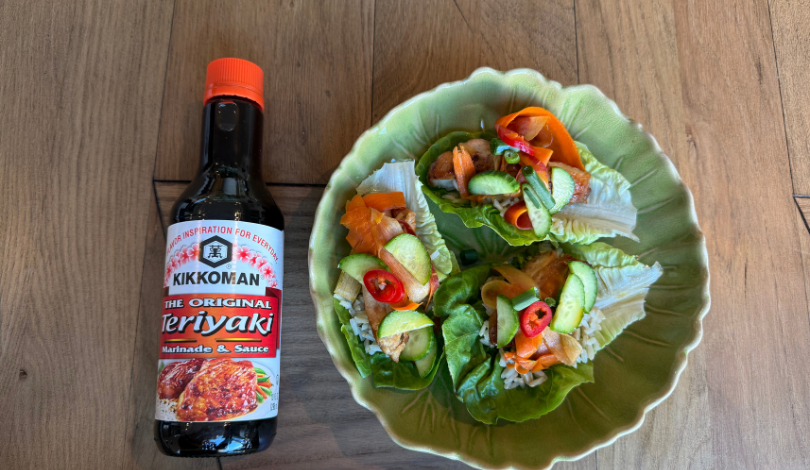 A bottle of Kikkoman Teriyaki sauce is next to a green bowl filled with lettuce wraps topped with vegetables and red chili slices on a wooden surface.
