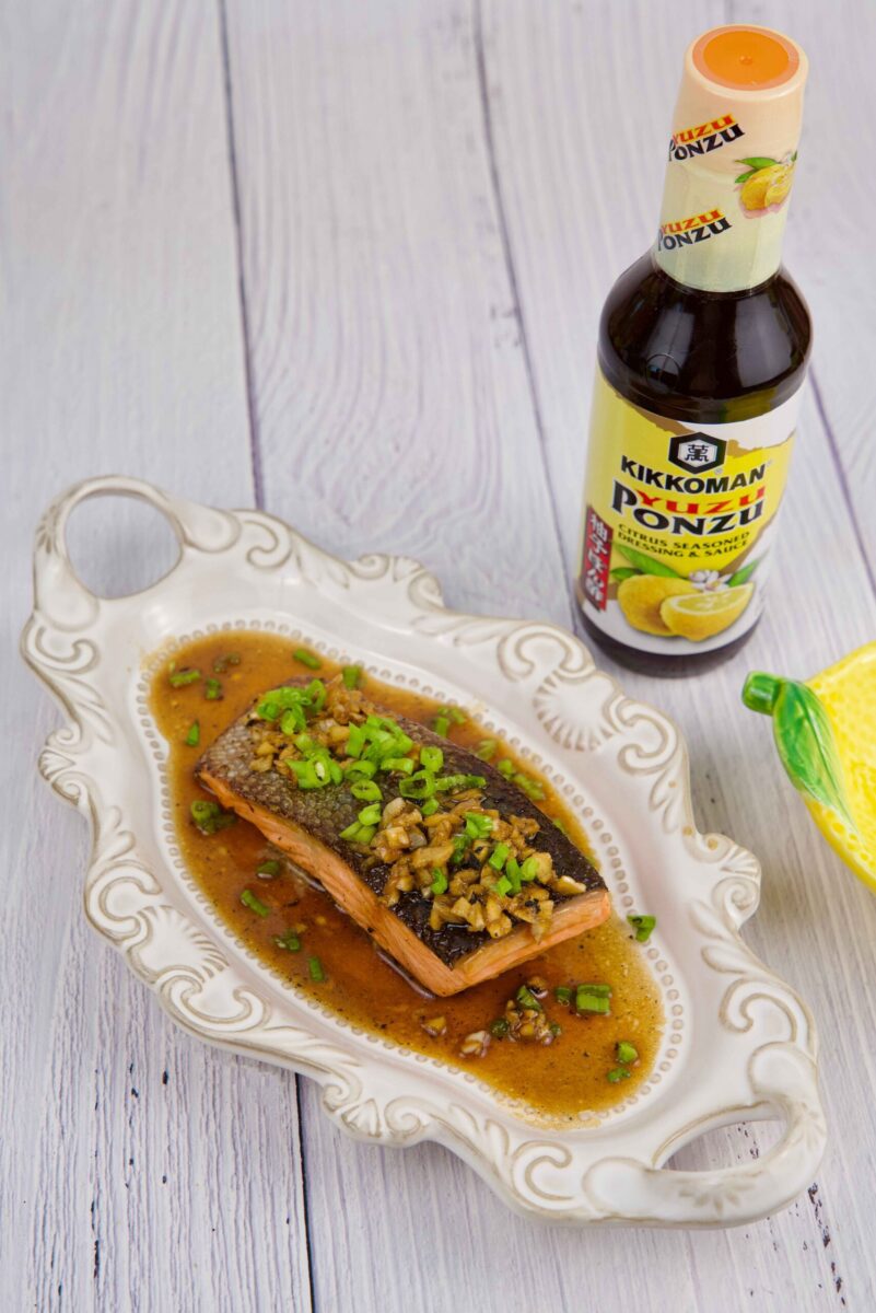 A cooked salmon fillet topped with chopped green onions and sauce is served on a decorative plate next to a bottle of Kikkoman Ponzu sauce.