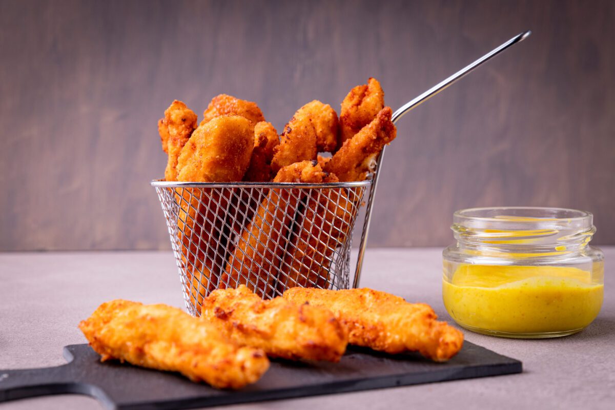 Fried chicken tenders in a metal basket with more tenders on a board and a small jar of yellow dipping sauce beside them.