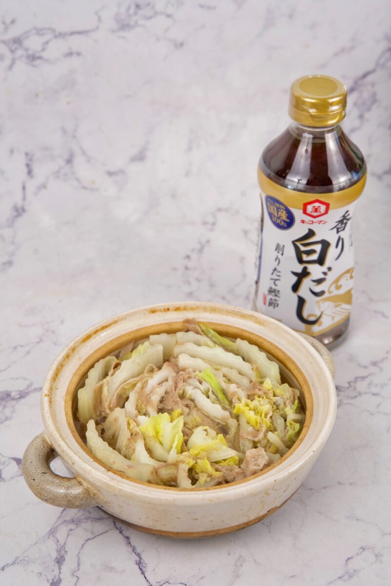 A ceramic pot filled with a layered cabbage and meat dish sits in front of a bottle of Japanese soup base on a light marble surface.
