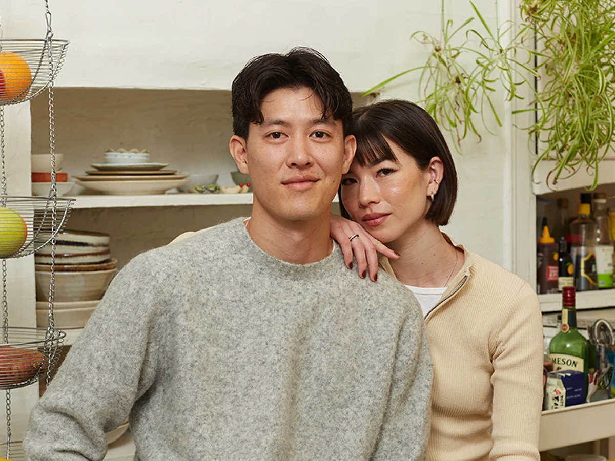 A man and a woman pose together in a kitchen, with shelves of dishes, fruit, and plants visible in the background.