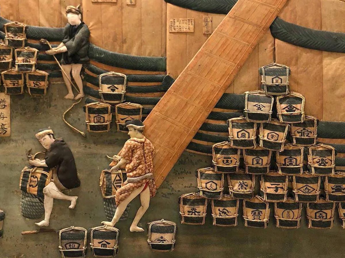 Three figures handle stacked barrels near a wooden ramp, with more barrels bearing Japanese writing arranged in the background. The setting appears historical and indoors.
