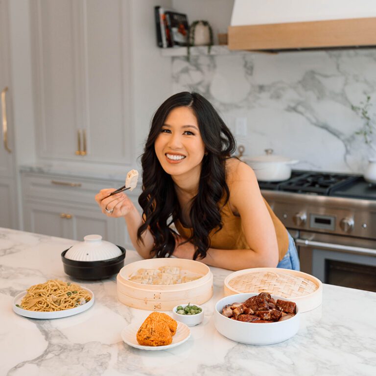 A woman sits at a kitchen counter, smiling while holding a dumpling. Various Asian dishes, including noodles, dumplings, and meats, are arranged in front of her.